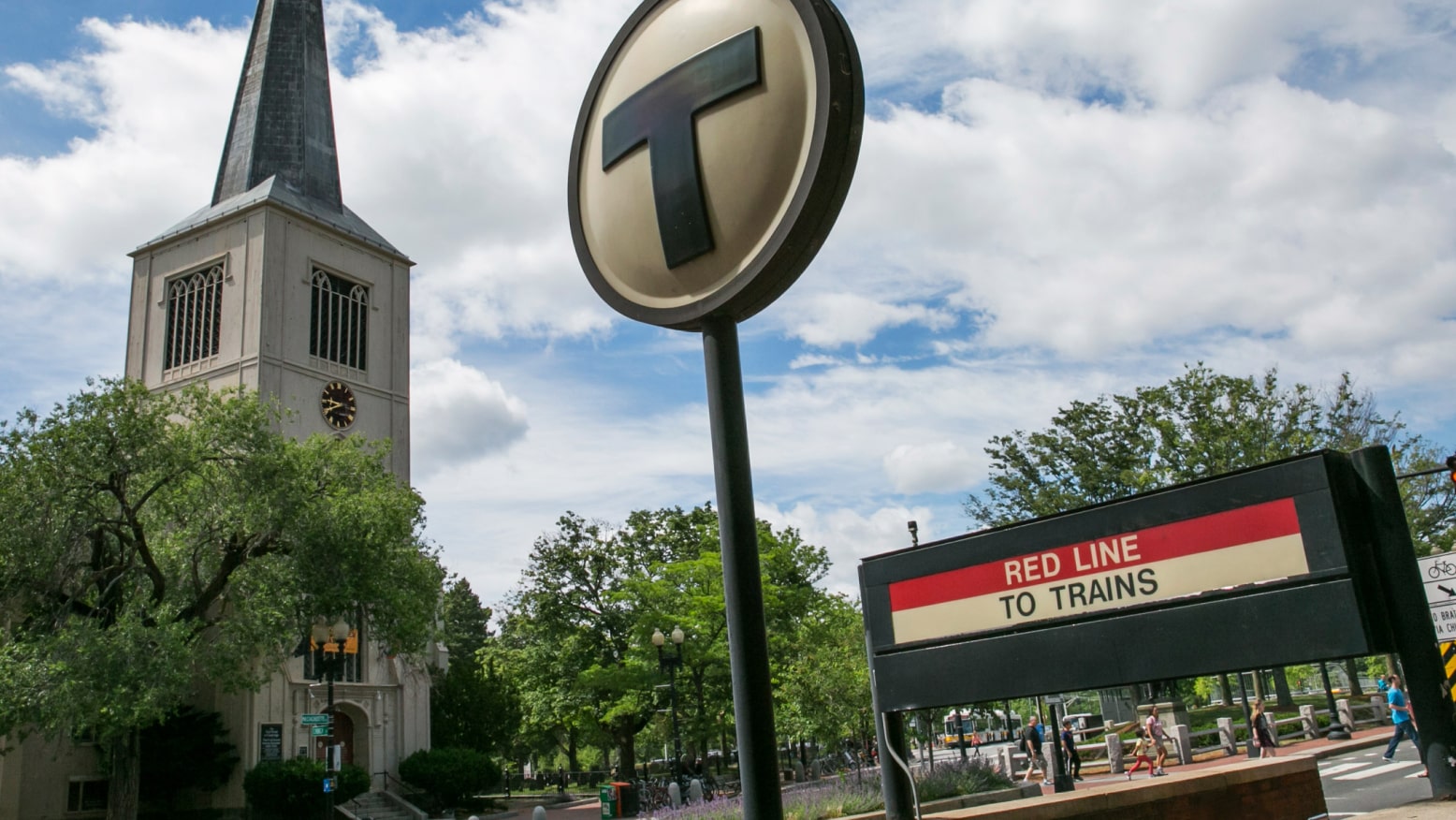 A train stop sign on a street in Cambridge, MA stands out against a blue sky with bright white clouds. A historic church steeple rises from the background.