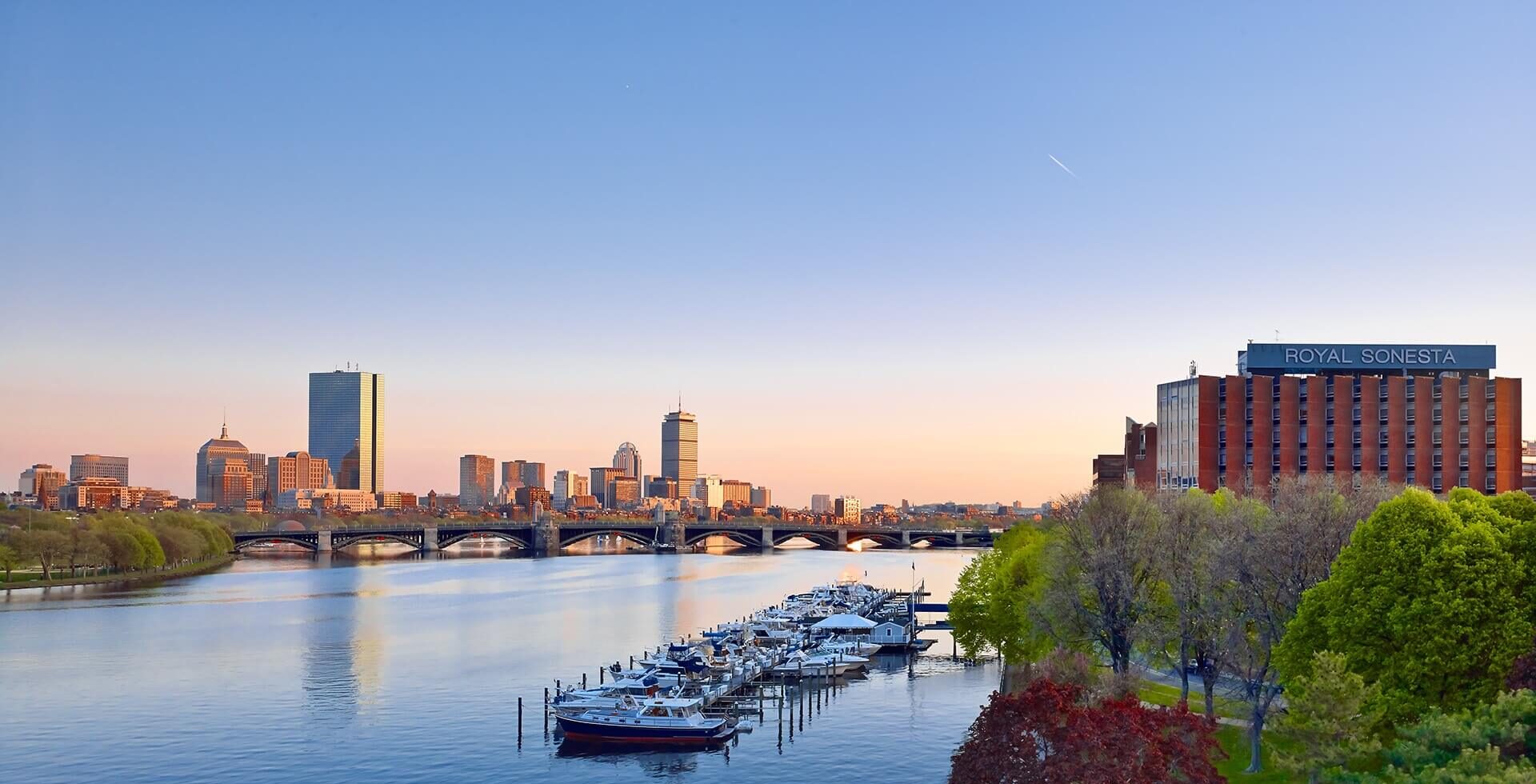 A tranquil view of the Charles River in Boston at sunset as seen from Cambridge MA, with boats docked at a marina in the foreground and the city skyline in the background. The Longfellow Bridge spans the river, and the Royal Sonesta Hotel is visible on the right surrounded by trees with spring foliage. The sky is clear with a soft gradient from blue to warm hues.