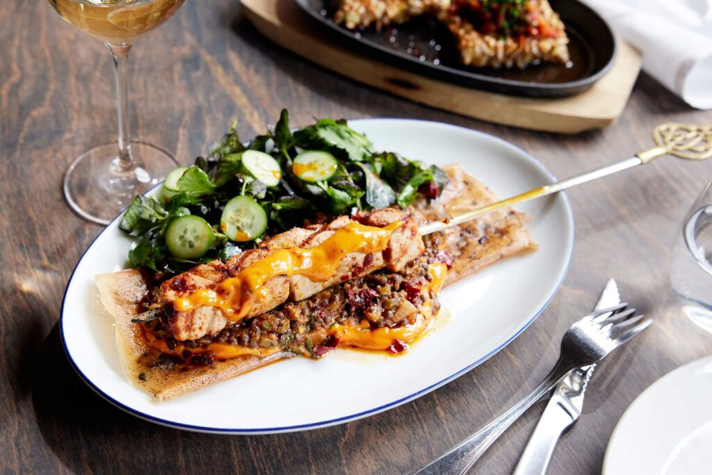 A plated gourmet meal featuring grilled chicken skewers drizzled with a vibrant orange sauce, served over a bed of savory lentils and flatbread at an upscale eatery in Cambridge. The dish is accompanied by a fresh mixed green salad with cucumber slices. A glass of white wine sits nearby on a wooden table, along with a fork and knife.