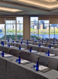 A well-lit conference room at a business-friendly hotel like the Boston Marriott Cambridge. It's set up classroom-style with rows of tables and chairs, each place equipped with a notepad, pen, glass, and blue water bottle. Large windows offer a scenic view of a river, city buildings, and trees outside, allowing natural light to brighten the room.