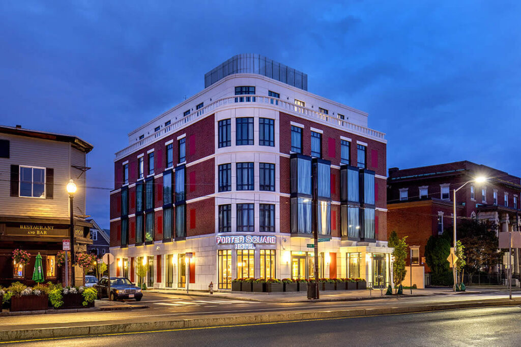 An evening view of the Porter Square Hotel in Cambridge MA, a modern red-brick and white-trim building located on a city corner. Warm lights glow from the lobby windows, and the sign “Porter Square Hotel” is clearly visible above the entrance. Nearby, a cozy restaurant and bar with outdoor seating adds charm to the urban setting under a deep blue sky.