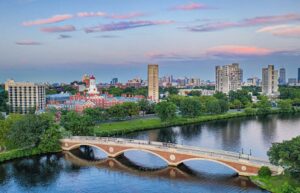 A scenic aerial view of the Charles River in Cambridge, Massachusetts, featuring a historic arched bridge with pedestrians crossing. In the background, Harvard University’s red-domed buildings and several tall residential towers are surrounded by greenery. The Boston skyline is visible in the distance under a colorful sky at sunset.