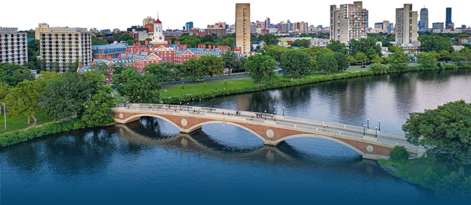 A late afternoon aerial view of Anderson Memorial bridge connecting two verdant green banks while the Cambridge MA skyline stands in the background.