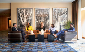 Five professionally dressed individuals sit comfortably in a modern lounge area, engaged in friendly conversation. Behind them are three large monochrome tree-themed artworks on a beige wall, and natural light pours in through a nearby window with brown curtains.