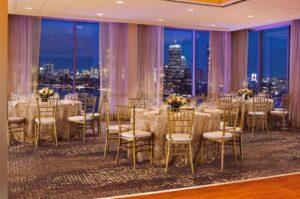 An elegant banquet hall with round tables covered in gold-patterned linens and surrounded by gold chairs, set for a formal dinner. Large windows offer a stunning nighttime view of a city skyline with illuminated skyscrapers and the iconic CITGO sign.