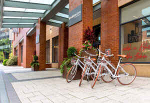 Exterior view of the Residence Inn by Marriott with three white rental bicycles parked under a glass canopy, surrounded by brick walls, potted plants, and a neon sign in the adjacent window reading ‘catch a Buzz like Aldrin