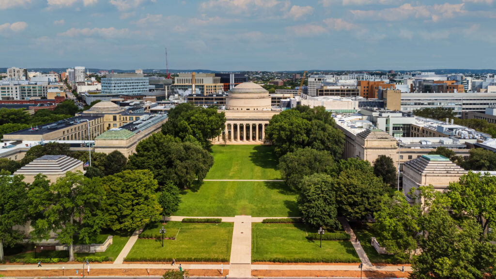 Aerial view of the Massachusetts Institute of Technology (MIT) campus, featuring the iconic Great Dome and expansive Killian Court surrounded by symmetrical neoclassical buildings and lush green trees, with the city of Cambridge visible in the background.