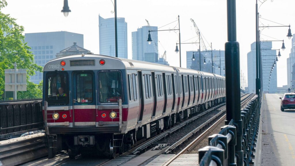 A red and silver subway train travels across a bridge into the city with modern skyscrapers in the background, passing street lamps and running parallel to a road with a red car and a few pedestrians