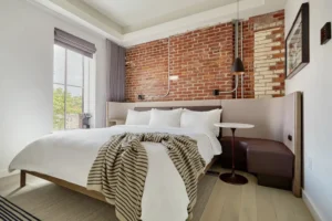 Modern hotel bedroom with white bed, exposed brick accent wall, large window, and minimalist furniture.